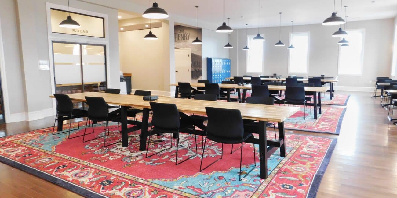 Spacious modern office with long wooden tables, black chairs, pendant lights, colorful rugs on wood floor, and glass-walled rooms.