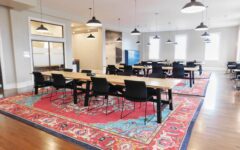 Spacious modern office with long wooden tables, black chairs, pendant lights, colorful rugs on wood floor, and glass-walled rooms.