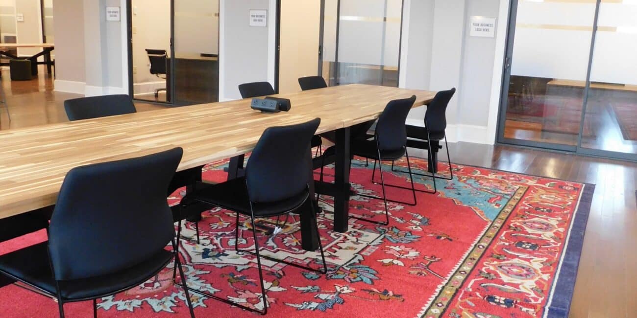 A modern conference room with a long wooden table, black chairs, hanging lights, colorful rug, and glass doors labeled Suite A-1 to A-3.