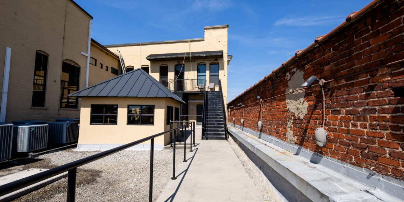A paved walkway with black railing leads to a yellow two-story building with exterior stairs beside a brick wall and AC units.