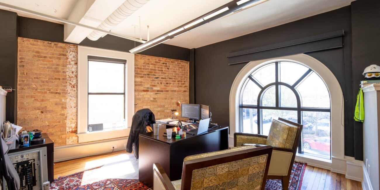Modern office with exposed brick, arched window, patterned chairs, desk and computer, colorful rug, wood floor, bright daylight.