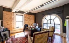 Modern office with exposed brick, arched window, patterned chairs, desk and computer, colorful rug, wood floor, bright daylight.