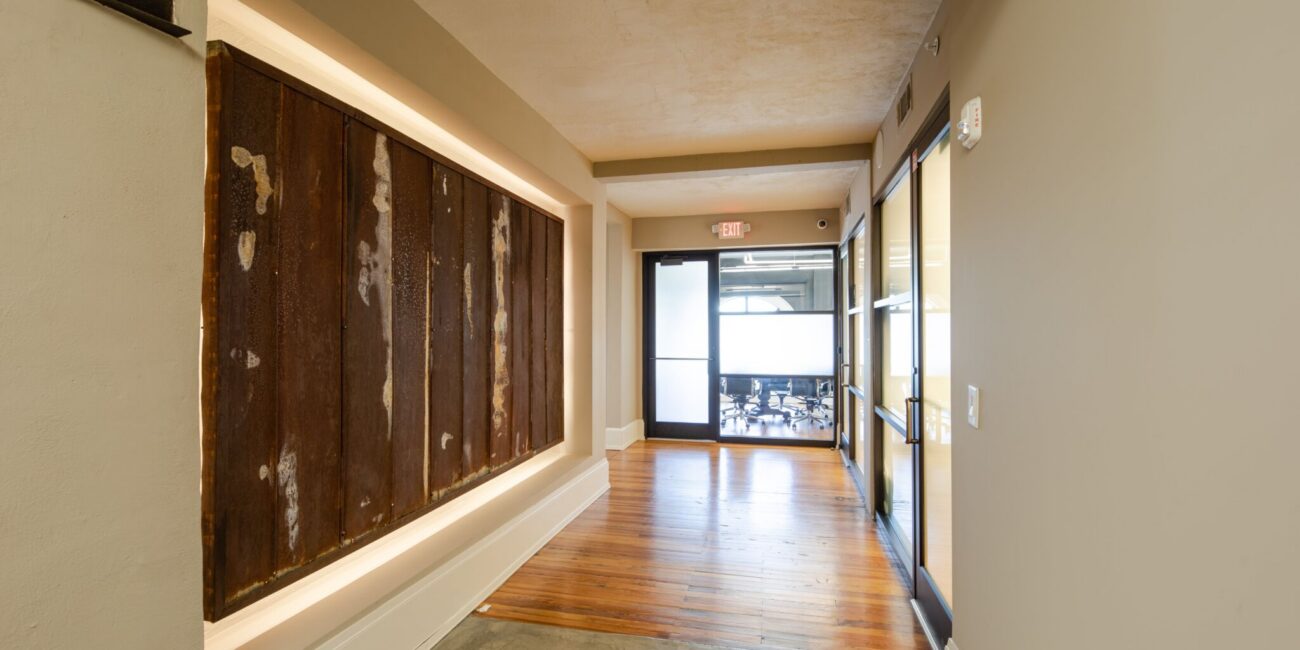 A hallway with beige walls, wood and metal paneling on the left, wood floor, glass double doors, and a visible conference room beyond.