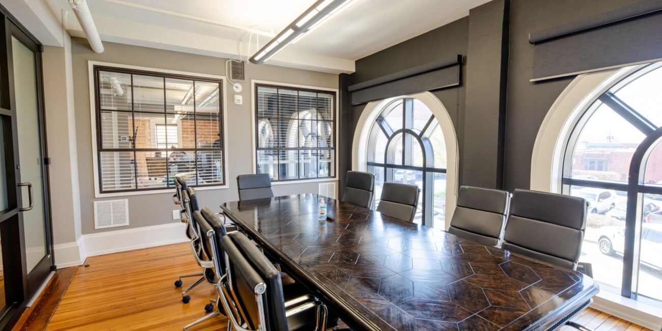 Modern conference room with dark wood table, black swivel chairs, hardwood floors, arched windows, and hand sanitizer on table.
