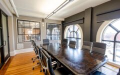 Modern conference room with dark wood table, black swivel chairs, hardwood floors, arched windows, and hand sanitizer on table.
