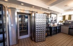 A restaurant interior with a large open metallic vault door, revealing wine storage and nearby glass cabinets of bottles and glasses.