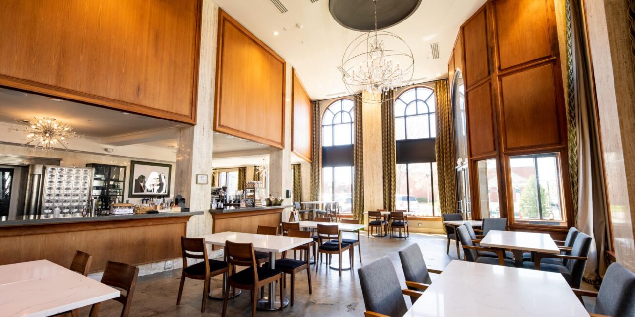 A spacious modern restaurant with high ceilings, arched windows, chandeliers, wood panels, marble tables, and a bar in back.