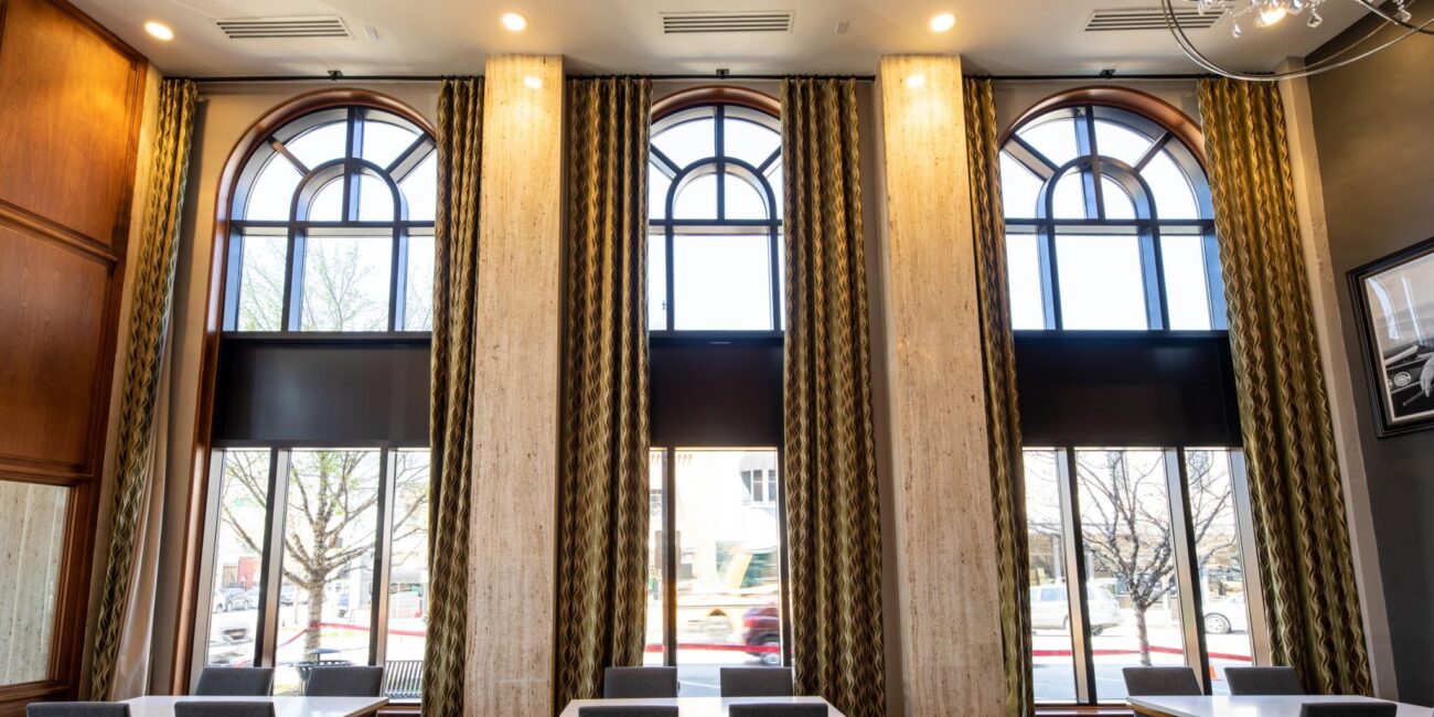 Spacious dining area with tall arched windows, patterned curtains, tables, and chairs. Trees and cars visible outside.