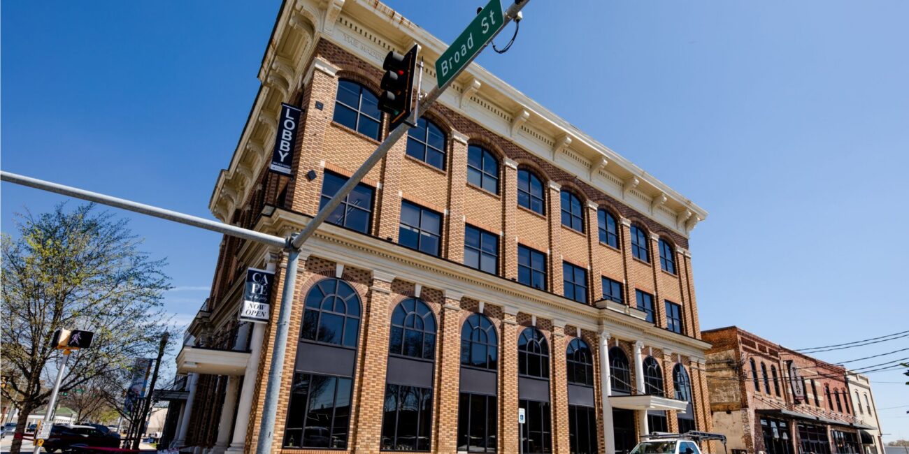 A three-story brick building on a street corner under a blue sky, arched windows, sign reading LOBBY. White truck and cones nearby; Broad St sign.