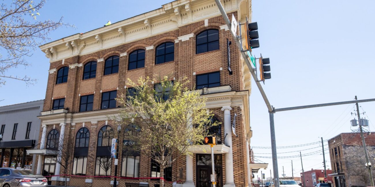 Three-story brick building with arched windows on a sunny corner, traffic lights and signs, parked cars, trees partly in front.