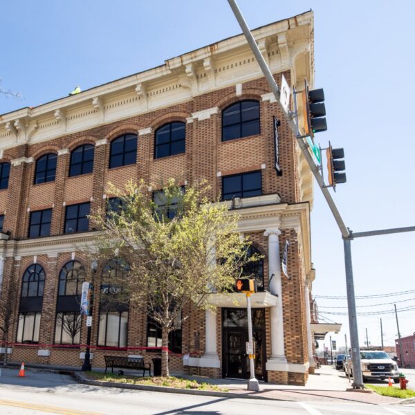 Three-story brick building with arched windows on a sunny corner, traffic lights and signs, parked cars, trees partly in front.