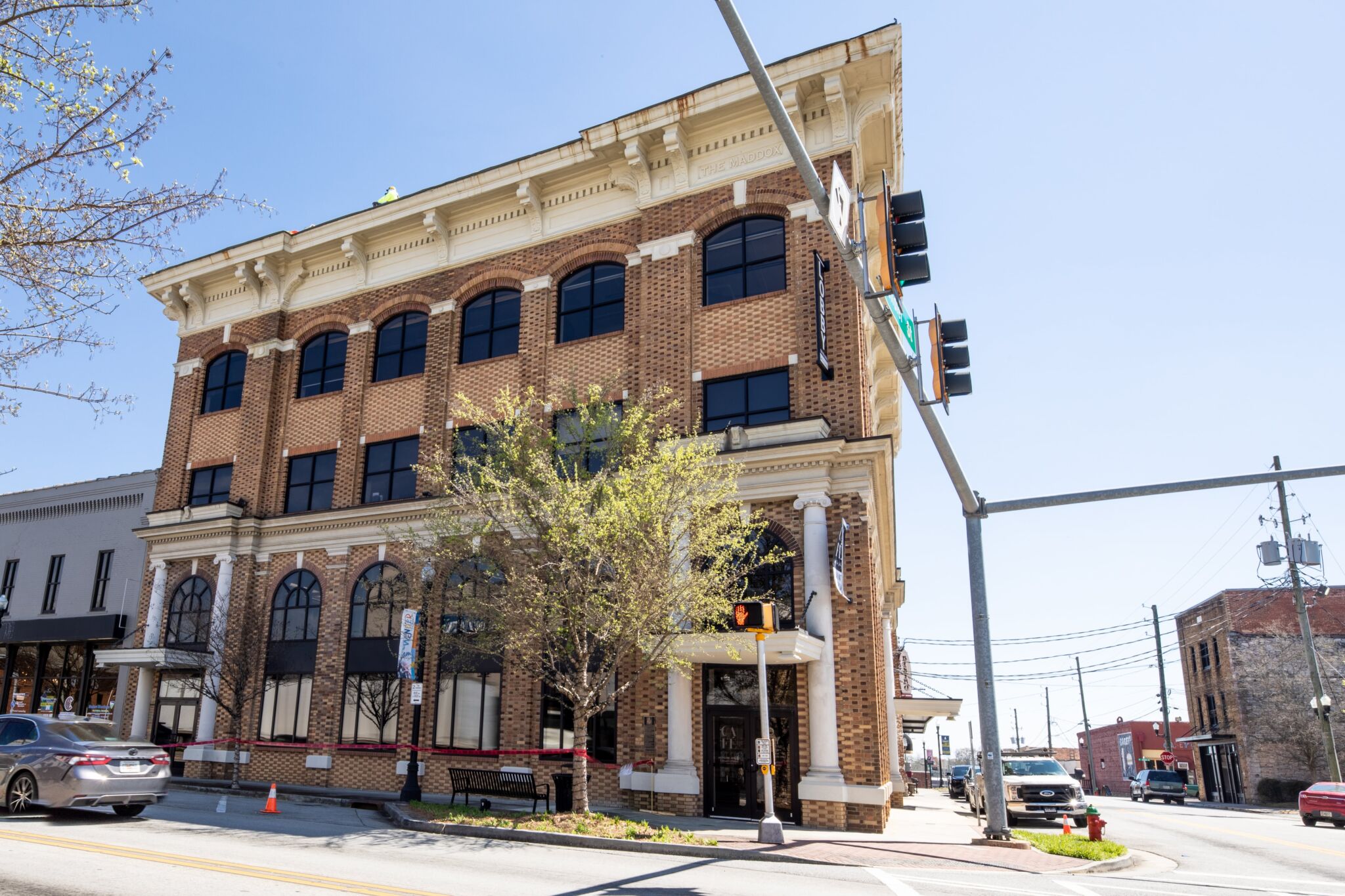 Three-story brick building with arched windows on a sunny corner, traffic lights and signs, parked cars, trees partly in front.