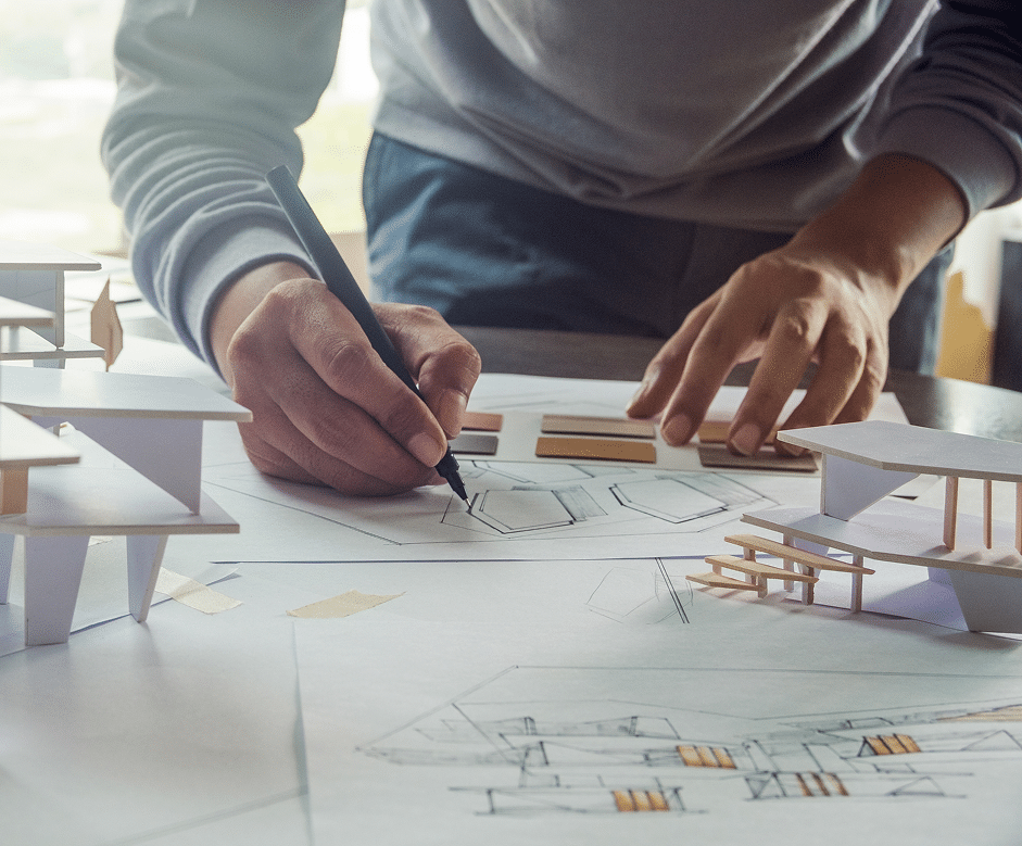 A person sketches architectural designs on paper amid model buildings and drafting tools, indicating work on a construction project.