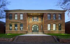 A historic two-story red brick building with Alexander written on the front, arched doorway, large windows, and steps, lit in evening.