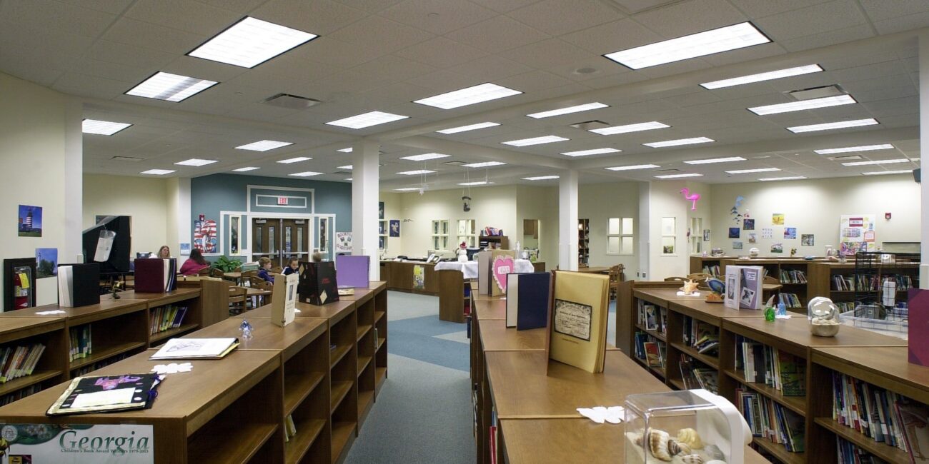 A spacious, well-lit library with wooden bookshelves, books, displays, and educational materials. Fluorescent lights hang overhead.