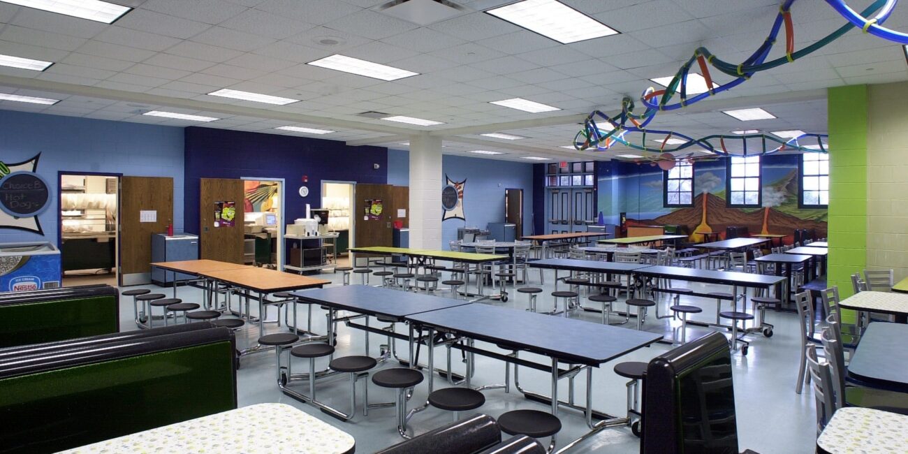 A brightly lit school cafeteria with long tables, round stools, colorful streamers, mural walls, and an empty food service area.