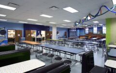 A brightly lit school cafeteria with long tables, round stools, colorful streamers, mural walls, and an empty food service area.