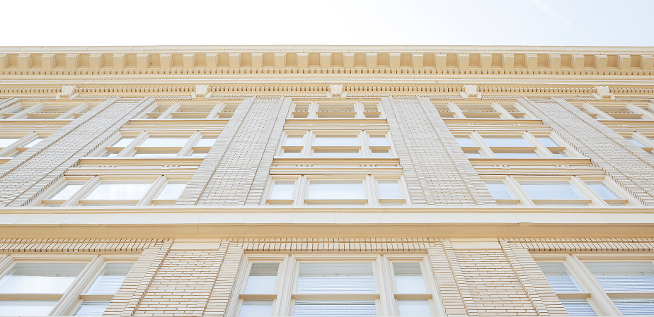 Looking up at a pale brick building featuring numerous large windows and ornate roof trim set against a bright sky.