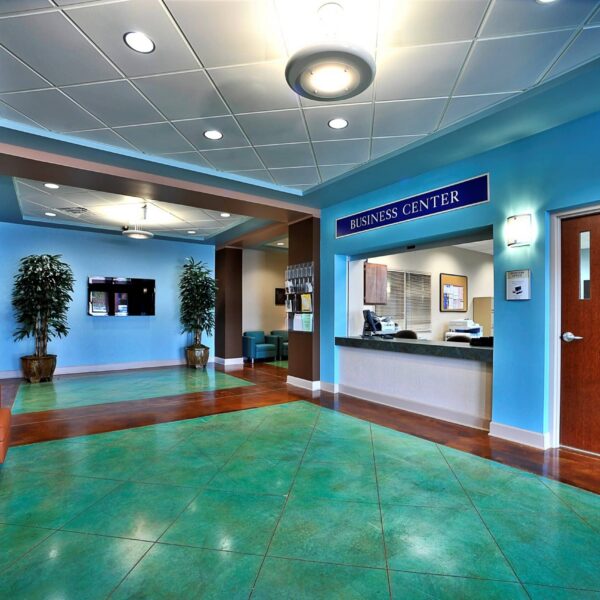 A modern business center lobby with teal floor tiles, blue walls, brown doors, plants, seating area, and a reception desk under a Business Center sign. Ceiling lights and wall decor create a welcoming atmosphere.