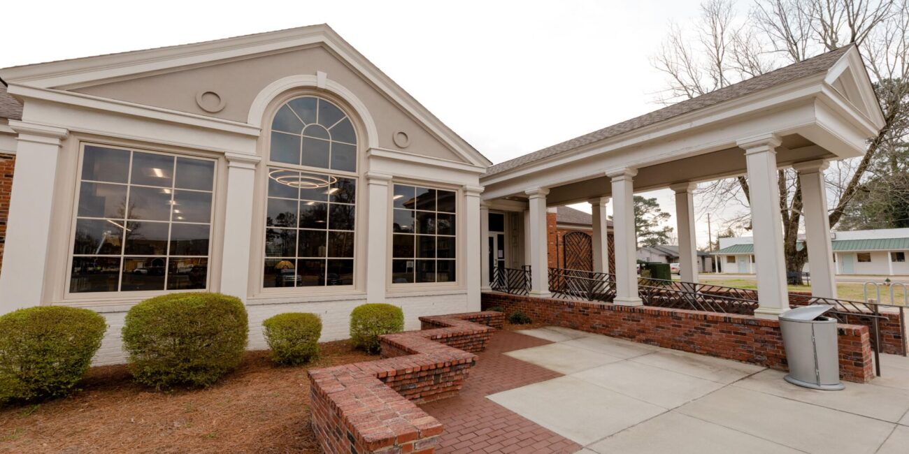 A single-story building features large arched windows and a covered entrance with white columns, bordered by bushes and a brick path.