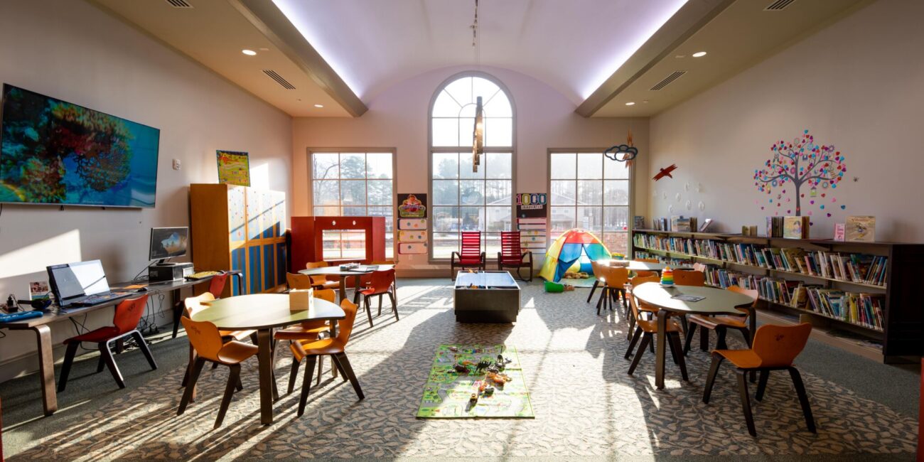 Bright, colorful children's playroom with small tables, chairs, bookshelves, a play tent, toy mat, wall mural, sunny arched windows.