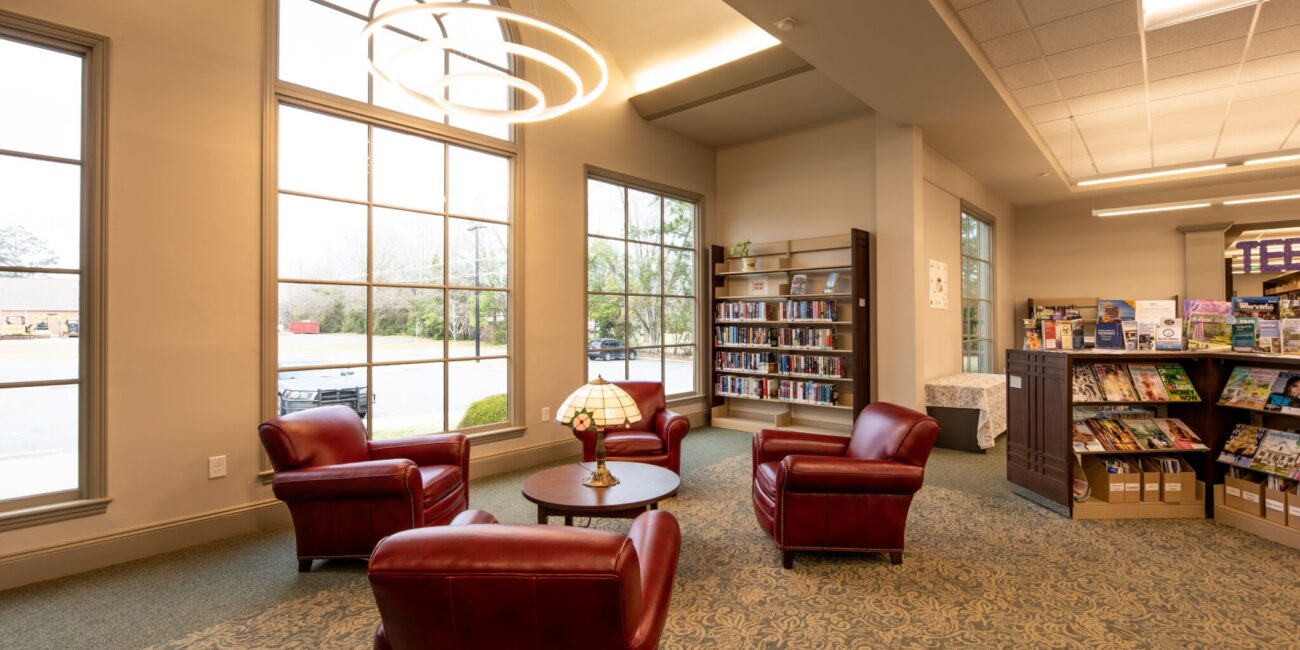 Cozy library reading area with four red leather chairs, a small table, large windows, bookshelves, magazine rack, and modern light fixture.