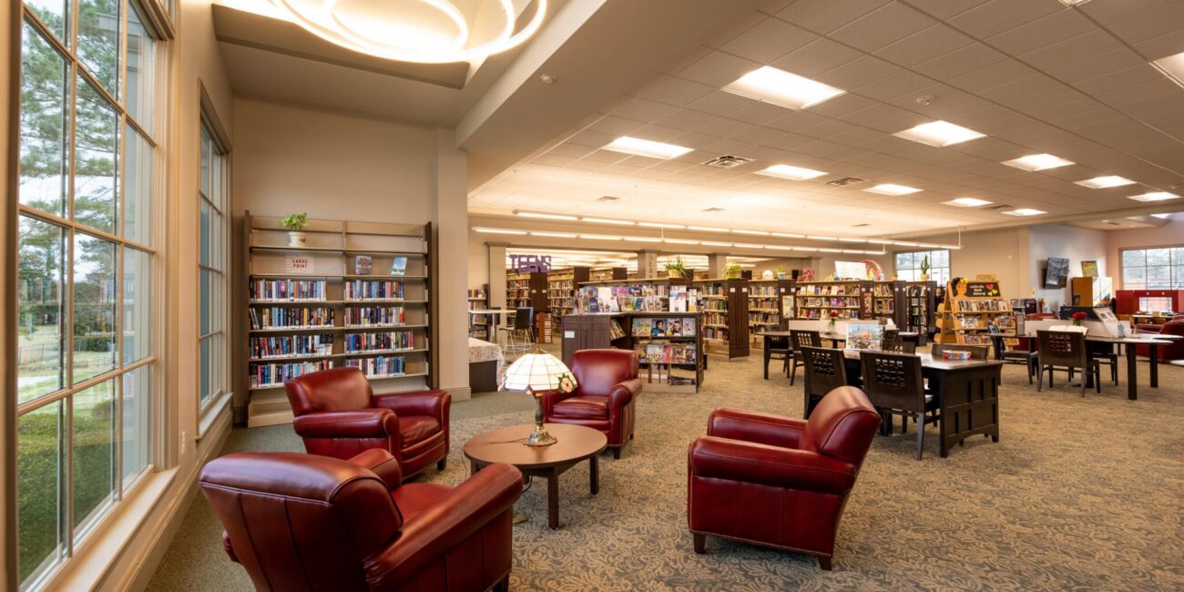 Spacious library with large windows, bookshelves, tables, and four red armchairs around a table under circular lights. Inviting space.