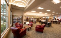 Spacious library with large windows, bookshelves, tables, and four red armchairs around a table under circular lights. Inviting space.