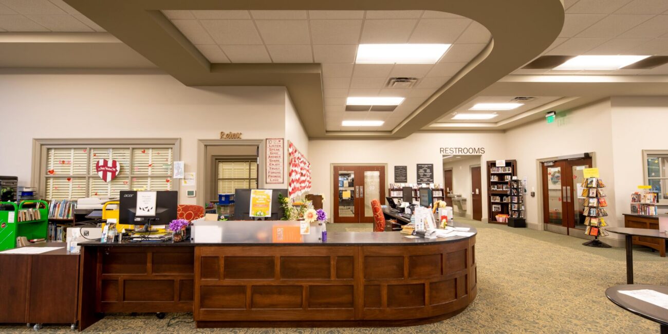 Wide view of a public library interior with a large wooden help desk, bookshelves, a Restrooms sign, and various signs and decor.
