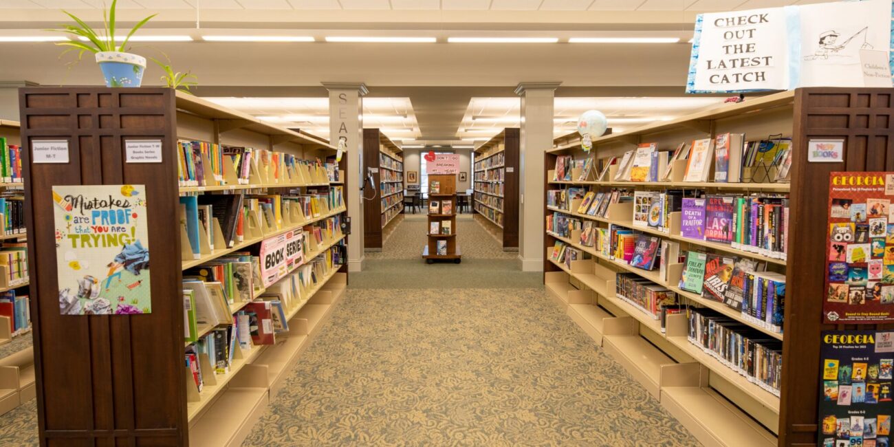 A brightly lit library with rows of bookshelves, colorful books, decorations, and a carpeted aisle leading to a sign reading CHECK OUT THE LATEST CATCH.