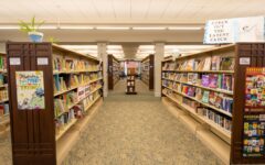 A brightly lit library with rows of bookshelves, colorful books, decorations, and a carpeted aisle leading to a sign reading CHECK OUT THE LATEST CATCH.