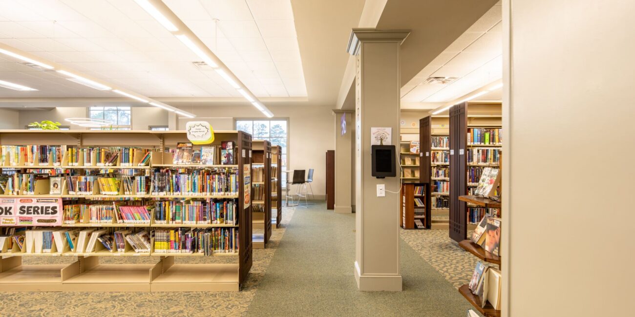 Bright library interior with bookshelves, a book series sign, carpeted floors, large windows, and a study area with chairs.