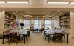 Spacious, well-lit library with round tables, dark chairs, bookshelves right, board games left, large back windows, circular ceiling lights.