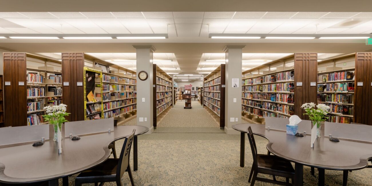 Spacious, bright library with bookshelves, round tables, chairs, flower vases, and tissues. Quiet, organized space for study or reading.