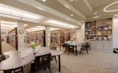 A spacious library features bookshelves, round tables with chairs and flower vases, plus a wall shelf of board games in bright light.