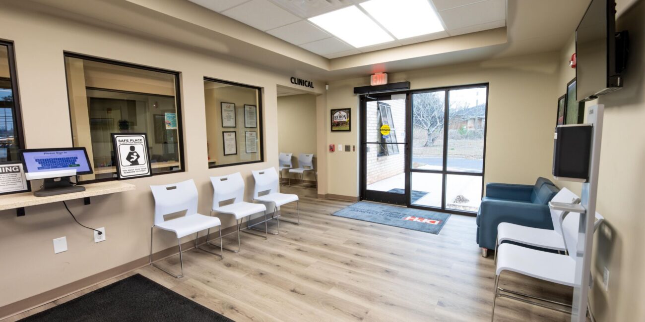 Clean, modern clinic waiting area with white chairs, a reception window, TV, glass doors, wood-look floors, and posted signs.