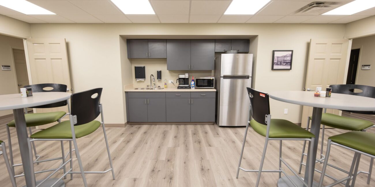 Modern break room with tall tables, gray kitchenette, microwave, coffee maker, stainless fridge, framed photo, light wood floors.