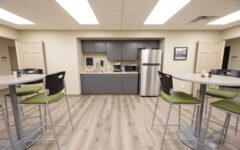 Modern break room with tall tables, gray kitchenette, microwave, coffee maker, stainless fridge, framed photo, light wood floors.