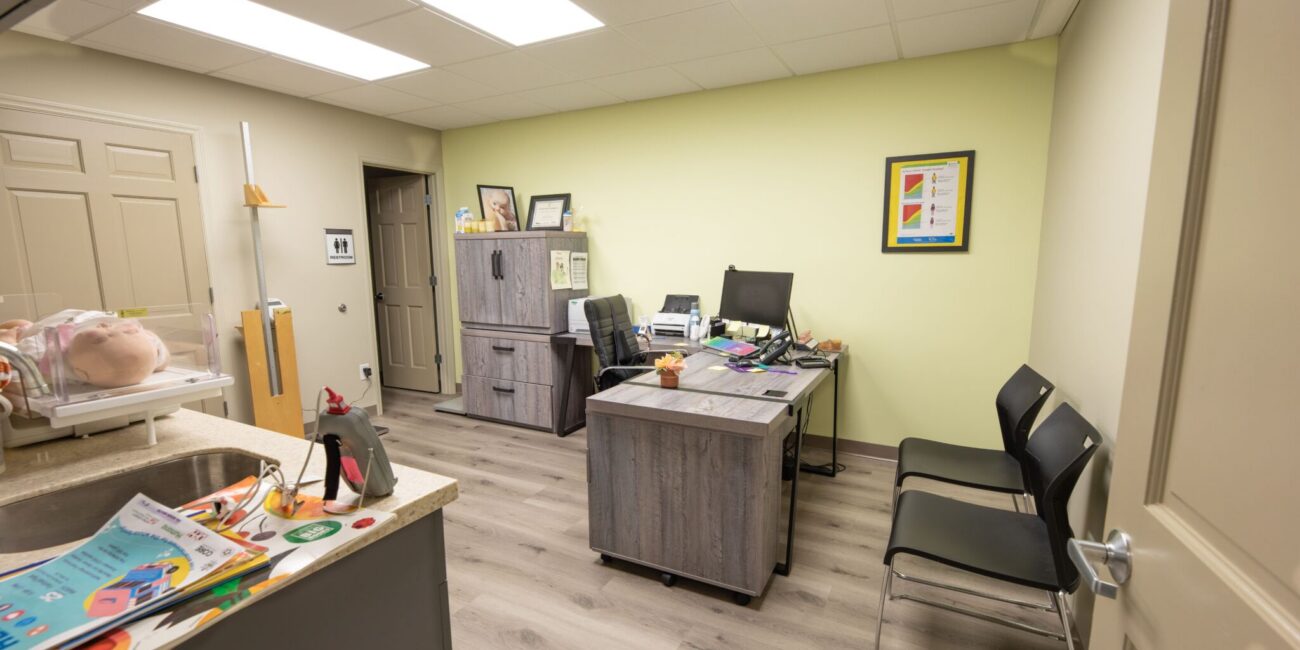 A tidy, modern medical office with two desks, visitor chairs, gray filing cabinet, charts and models, well-lit with ceiling lights.
