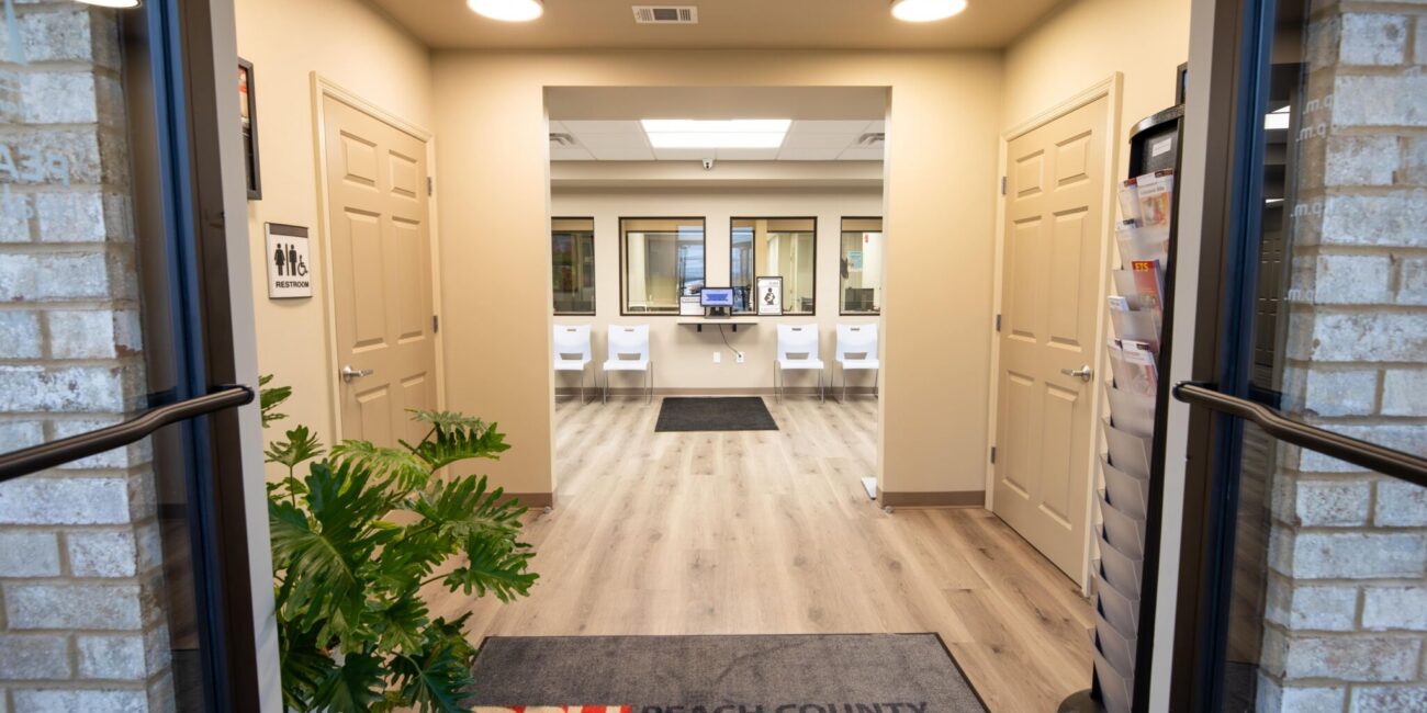 View through open double doors into a modern health office with wood floors, beige walls, a plant, reception desk, and Peach County Health Department mat.