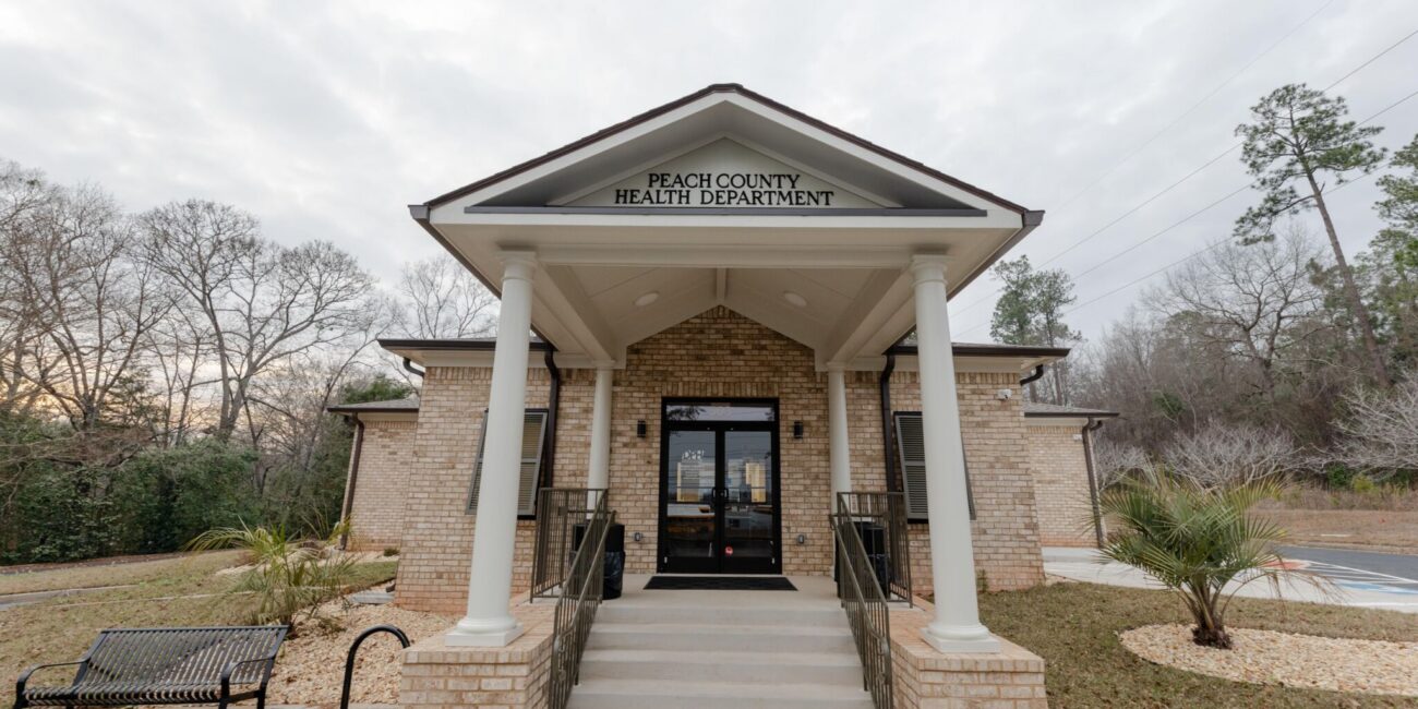 Entrance of Peach County Health Department with brick pillars, steps, glass doors, a left-side bench, and landscaped grounds with trees.