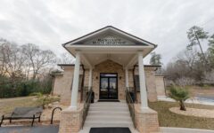 Entrance of Peach County Health Department with brick pillars, steps, glass doors, a left-side bench, and landscaped grounds with trees.