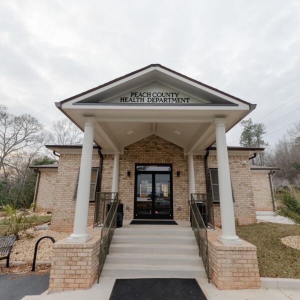 Entrance of Peach County Health Department with brick pillars, steps, glass doors, a left-side bench, and landscaped grounds with trees.