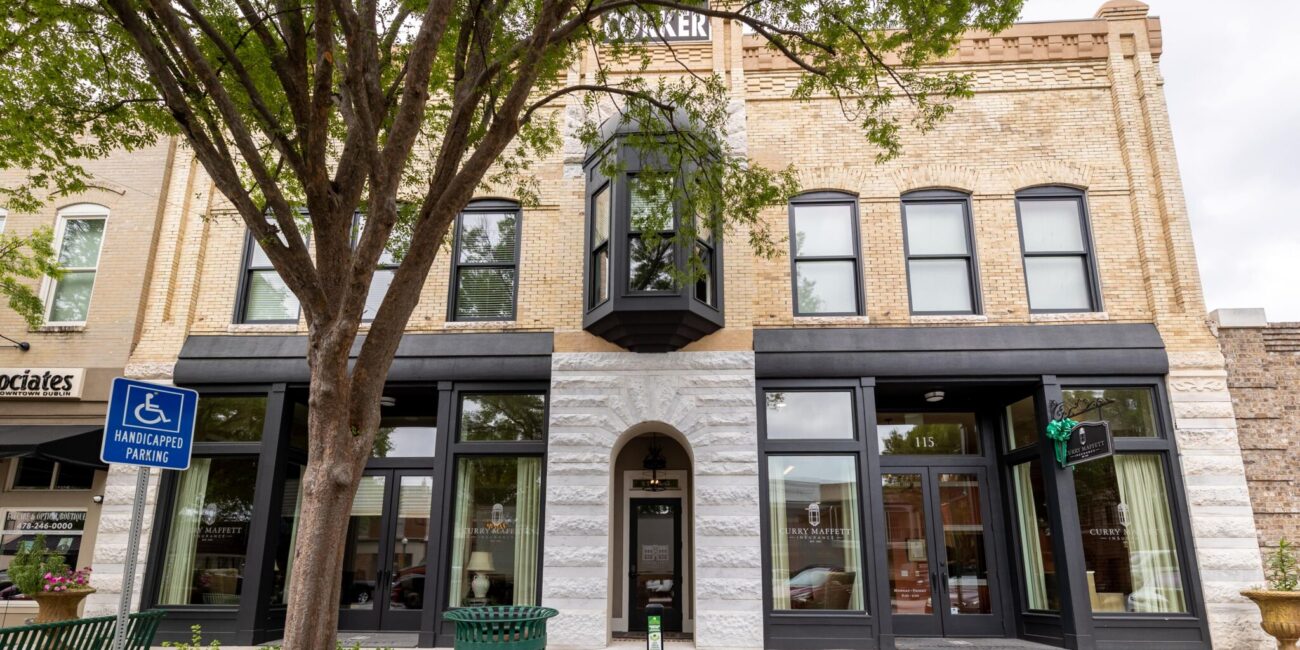 Two-story brick building with large arched and rectangular windows, a glass-door entrance, tree out front, and handicapped parking sign.