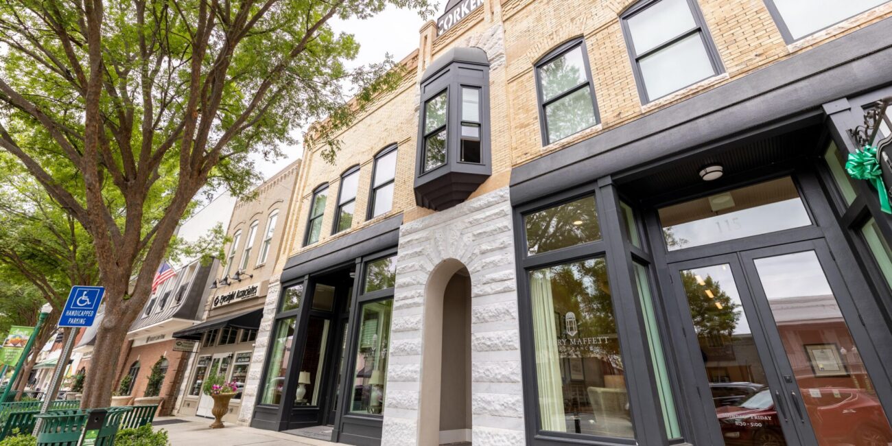 Street view of historic brick building with large windows, stone arch entry, black bay window, trees, green railings, and handicap sign.