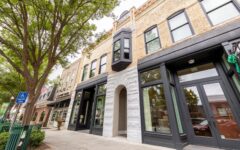Street view of historic brick building with large windows, stone arch entry, black bay window, trees, green railings, and handicap sign.