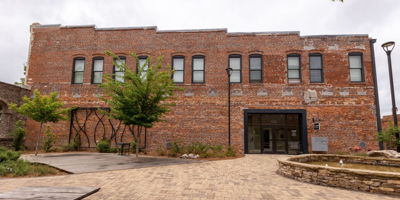 Two-story brick building with six upper windows, central entrance, courtyard with trees, benches, lamp post, fountain. Cloudy sky.