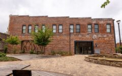 Two-story brick building with six upper windows, central entrance, courtyard with trees, benches, lamp post, fountain. Cloudy sky.