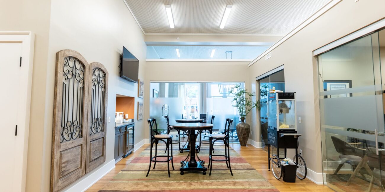 Modern office lounge featuring a round table, chairs on striped rug, decorative panels, kitchenette, glass rooms, and large windows.