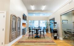 Modern office lounge featuring a round table, chairs on striped rug, decorative panels, kitchenette, glass rooms, and large windows.